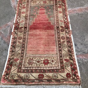 May include: A vintage Turkish prayer rug with a red, beige, and green color scheme. The rug has a traditional design with a central prayer niche and intricate floral patterns along the border.