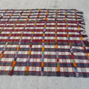 May include: A white and red checkered rug with colorful fringe. The rug has a woven texture and is made of wool.