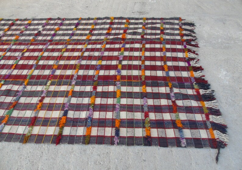 May include: A white and red checkered rug with colorful fringe. The rug has a woven texture and is made of wool.