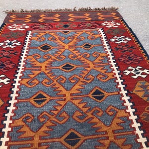 May include: A rectangular rug with a central blue field featuring orange geometric patterns. The rug has a red border with white and black geometric designs. The top edge has a brown fringe.