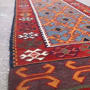 May include: A long, rectangular rug with a detailed geometric pattern. The rug features a central design in shades of blue and orange, framed by a red border with white and dark blue accents. The rug has a fringed edge.