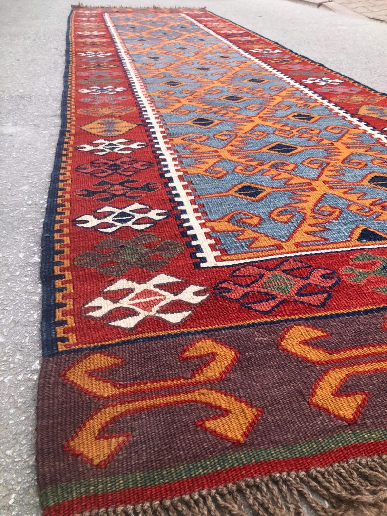 May include: A long, rectangular rug with a detailed geometric pattern. The rug features a central design in shades of blue and orange, framed by a red border with white and dark blue accents. The rug has a fringed edge.