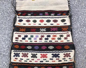 May include: Handwoven bag with a cream-colored background and colorful geometric patterns in black, red, orange, purple, and blue. The bag has a braided strap and a tassel detail.