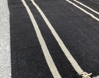 May include: A black and white striped rug with a woven texture. The rug features thick, vertical white stripes against a dark background. The edges have a fringe detail. This rug is suitable for indoor or outdoor use.