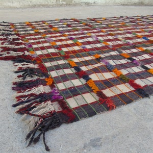 May include: A colorful woven rug with a checkered pattern and fringe. The rug is made of wool and has a variety of colors, including red, white, black, orange, and purple.