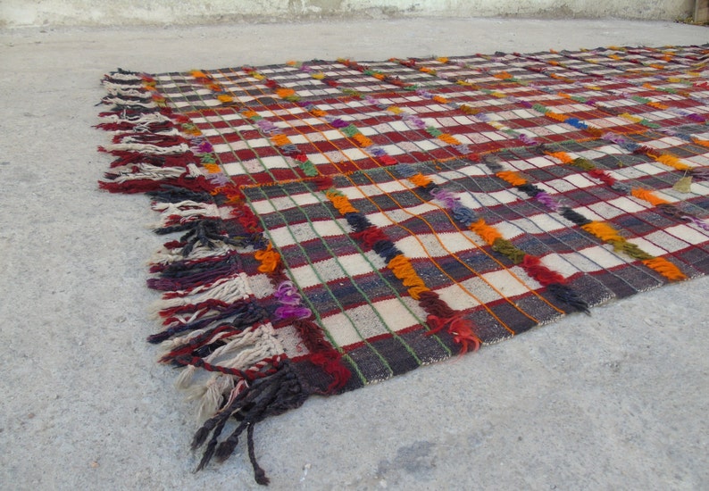 May include: A colorful woven rug with a checkered pattern and fringe. The rug is made of wool and has a variety of colors, including red, white, black, orange, and purple.