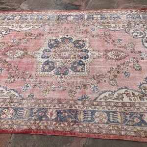 May include: A vintage rug with a faded pink background and intricate floral patterns in shades of blue, beige, and cream. The rug has a distressed look with a worn border.