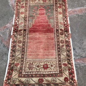 May include: A vintage Turkish prayer rug with a red, beige, and green geometric pattern. The rug has a fringe border and is in good condition.