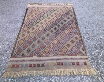May include: Rectangular rug with a geometric diamond pattern in shades of beige, red, blue, and yellow. The rug has a fringed edge and a border with repeating geometric shapes. The rug is made of woven material.