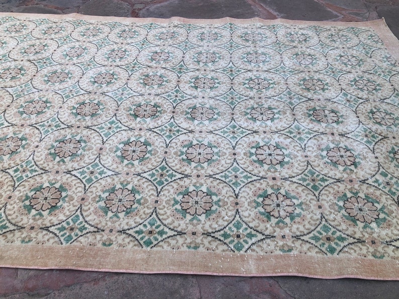 May include: A vintage rug with a repeating pattern of green, brown, and white floral motifs. The rug has a light beige background and a pink border.