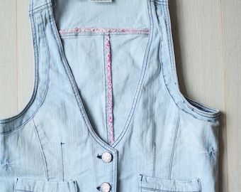 Op de afbeelding: Een lichtblauw denim vest met een roze en witte bloemenbies rond de halslijn. Het vest heeft twee zakken aan de voorkant en drie knopen in het midden.