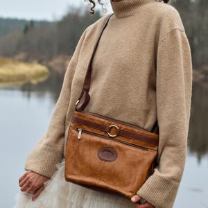 May include: A brown leather crossbody bag with a gold-tone buckle and zipper detail. The bag has a shoulder strap and a logo on the front. The person is wearing a beige sweater and a white skirt.