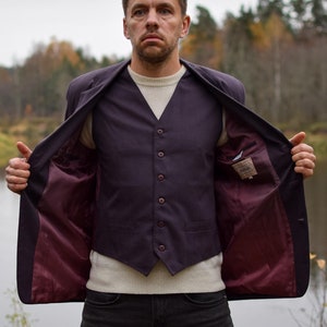 May include: A man wearing a dark purple vest and a dark purple jacket with a maroon lining, standing in front of a river with fall foliage in the background.