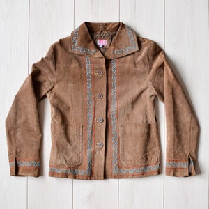 Op de afbeelding: Een bruin suède jack met blauwe en oranje borduurwerk rond de kraag, zakken en manchetten.