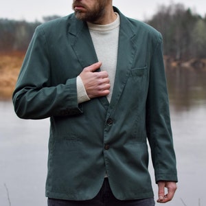 Könnte beinhalten: Ein Mann in einem dunkelgrünen Blazer über einem weißen Pullover, der an einem See steht. Er blickt nach links aus dem Bild.