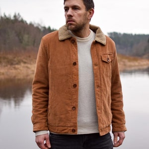 Könnte beinhalten: Ein Mann trägt eine braune Cordjacke mit Fleecefutter, einen weißen Pullover und eine blaue Jeans. Er steht vor einem Gewässer mit Bäumen im Hintergrund.