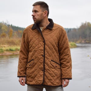 Könnte beinhalten: Braune Steppjacke mit schwarzem Kragen und Besatz. Die Jacke hat eine Knopfleiste und zwei Vordertaschen. Die Person steht in der Nähe eines Flusses mit Bäumen im Hintergrund. Die Jacke hat eine mittelbraune Farbe.