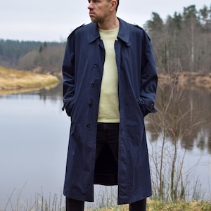 Könnte beinhalten: Ein marineblauer Trenchcoat mit Knopfleiste und Kragen. Der Mantel wird über einem hellgelben Hemd und einer schwarzen Hose getragen. Die Person steht in der Nähe eines Flusses.