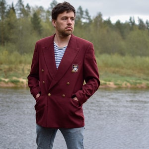 Puede incluir: Un hombre con un blazer de doble botonadura color granate con botones dorados y un pequeño emblema en el lado izquierdo. Está de pie frente a un río con un bosque en el fondo.