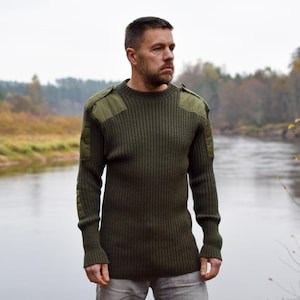 Może przedstawiać: Oliwkowozielony sweter z prążkowanej dzianiny ze wzmocnieniami na ramionach i łokciach. Sweter z długim rękawem ma okrągły dekolt i wojskowy design. Osoba stoi na zewnątrz w pobliżu rzeki.