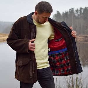 May include: A brown corduroy jacket with a navy collar and a red and black tartan lining. The jacket has a leather accent on the front and a navy lining. The person is wearing a pale yellow top and dark jeans.