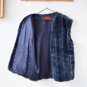 Könnte beinhalten: Eine marineblaue Kunstfellweste mit Satinfutter, die an einem schwarzen Kleiderbügel hängt. Die Weste hat einen Knopfverschluss und ein Etikett mit der Aufschrift "uhle". Die West ist vor einer weißen Wand ausgestellt.