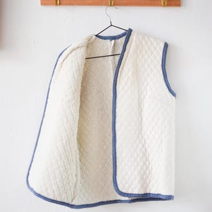 Könnte beinhalten: Eine cremefarbene, gesteppte Weste mit blauem Besatz hängt an einem schwarzen Kleiderbügel. Die Weste hat ein Rautenmuster auf der Außenseite und ein weiches, flauschiges Innenfutter. Sie hängt an einer Wand mit einer Holzgarderobe und Messing-Haken.