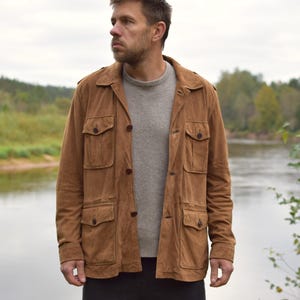 Könnte beinhalten: Eine braune Wildlederjacke mit vier Vordertaschen und Knopfverschlüssen. Die Jacke wird über einem grauen Pullover getragen. Die Person steht in der Nähe eines Flusses mit Bäumen im Hintergrund.