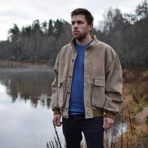 Puede incluir: Un hombre con una chaqueta de gamuza marrón está de pie junto a un lago con un bosque en el fondo. Lleva una camiseta azul y unos vaqueros azules.