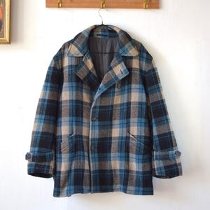 Könnte beinhalten: Ein Vintage-Mantel mit Karomuster in Blau-, Schwarz- und Beigetönen hängt an einem schwarzen Kleiderbügel. Der Mantel hat einen klassischen Kragen, einen Knopfverschluss und zwei Vordertaschen. Der Mantel hängt an einer Holzgarderobe mit Messinghaken.