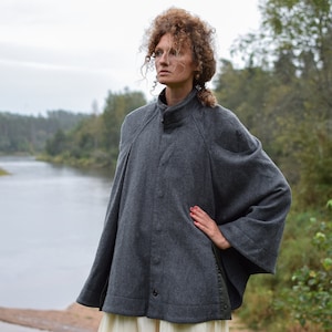 May include: A gray wool cape with a high collar and a button closure. The cape has a relaxed fit and is worn by a person standing in front of a river and trees.