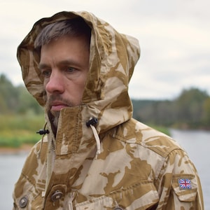 Könnte beinhalten: Eine Kapuzenjacke im Wüsten-Tarnmuster in Braun- und Beigetönen. Auf dem Ärmel befindet sich ein britischer Flaggenaufnäher. Die Person trägt die Jacke im Freien.