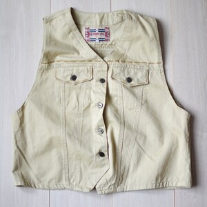 Può includere: Un giubbotto di jeans beige chiaro con chiusura a bottoni e due tasche. Il giubbotto ha un'etichetta che recita "The Original".