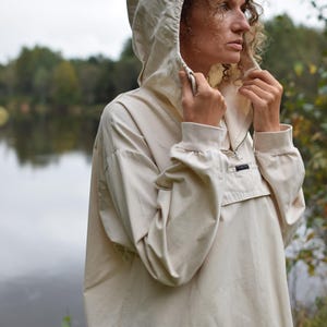 Puede incluir: Una chaqueta tipo pullover de color beige con capucha, cierre de media cremallera en el cuello, mangas largas con puños elásticos y un bolsillo frontal con solapa. La prenda está confeccionada en un tejido liso de color sólido.