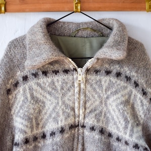Könnte beinhalten: Ein grauer Reißverschluss-Pullover mit einem geometrischen Muster in Weiß und Dunkelbraun. Der Pullover hat einen breiten Kragen und hängt an einem schwarzen Kleiderbügel. Der Hintergrund ist eine weiße Wand.