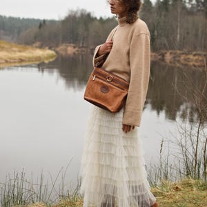 May include: A person wearing a tan sweater and a long white tiered skirt, carrying a brown leather shoulder bag with a gold-colored ring detail. The background features a river and trees.