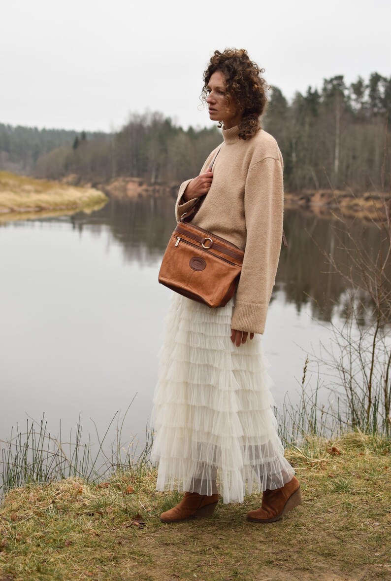 May include: A person wearing a tan sweater and a long white tiered skirt, carrying a brown leather shoulder bag with a gold-colored ring detail. The background features a river and trees.