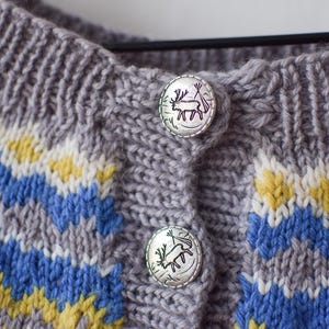 May include: Close-up of a gray knitted cardigan with blue, yellow, and white geometric patterns. The cardigan has two silver buttons with a deer design. The knitwear is a cozy, handmade garment.