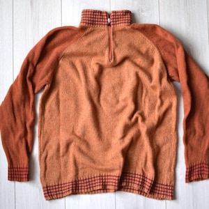 Puede incluir: Un suéter marrón con cuello alto y cremallera. El suéter tiene un patrón de cuadros rojos y blancos alrededor del cuello y los puños.