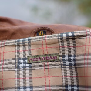 以下が含まれることがあります： 茶色の革のジャケットのクローズアップ。裏地はチェック柄。ジャケットにはXLサイズのタグと「rock n blue」のラベルが付いています。裏地は、赤と黒のチェック柄のタンです。