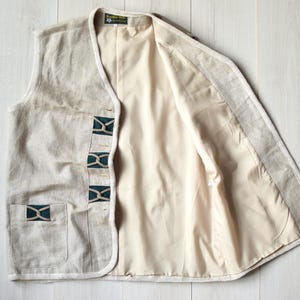 Könnte beinhalten: Eine beige Leinenweste mit grünem und braunem Musterbesatz. Die Weste hat eine Tasche auf der linken Seite und einen Knopfverschluss.