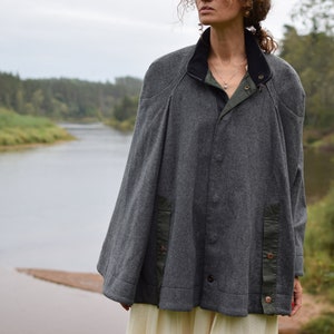 May include: A gray wool cape with a black collar and green accents. The cape has a button closure and is worn over a white dress.
