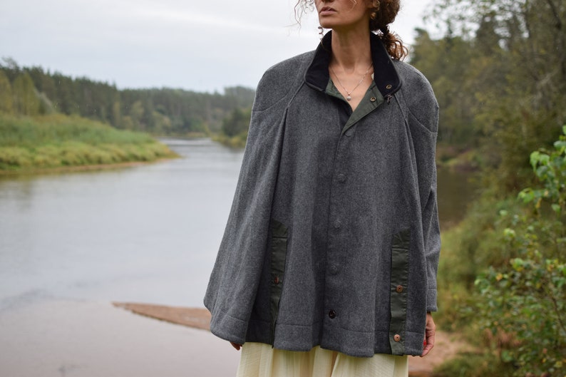 May include: A gray wool cape with a black collar and green accents. The cape has a button closure and is worn over a white dress.