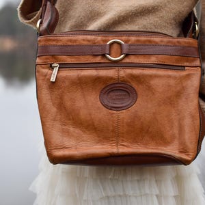 May include: A brown leather crossbody bag with a rectangular shape. The bag features a front zipper pocket, a circular logo, and a gold-colored ring detail. The bag is worn over a tan sweater and white skirt.