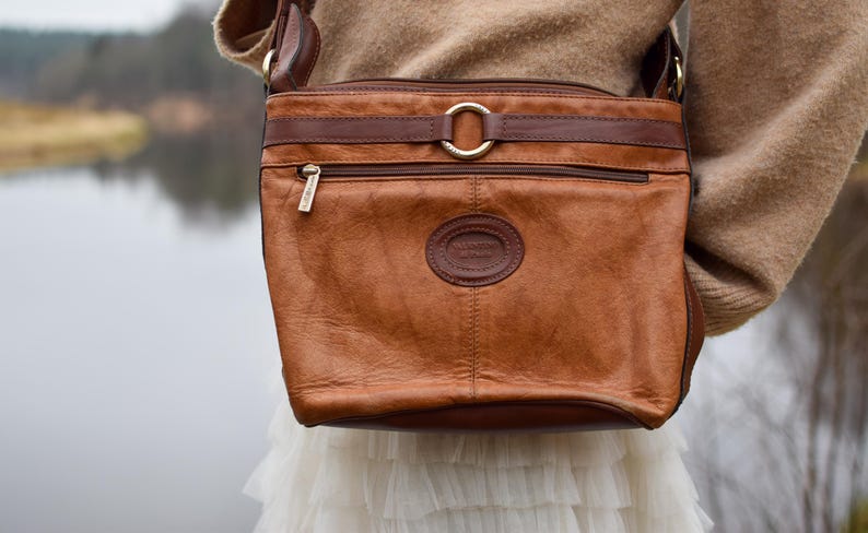 May include: A brown leather crossbody bag with a rectangular shape. The bag features a front zipper pocket, a circular logo, and a gold-colored ring detail. The bag is worn over a tan sweater and white skirt.