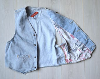 Op de afbeelding: Een lichtblauw denim vest met een rood label en een gepatenteerde voering. Het vest heeft een knoopsluiting en twee zakken.