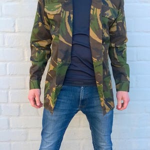 May include: A man wearing a camouflage jacket over a dark blue shirt and blue jeans. He is standing in front of a white brick wall.