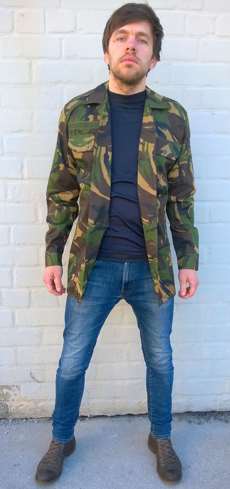 May include: A man wearing a camouflage jacket over a dark blue shirt and blue jeans. He is standing in front of a white brick wall.