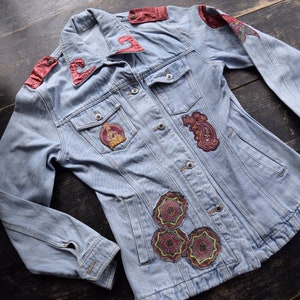 May include: Light blue denim jacket with decorative patches. The jacket has red velvet accents on the shoulders and collar. Intricate paisley and floral patches adorn the front, adding a unique, vintage flair to this garment.