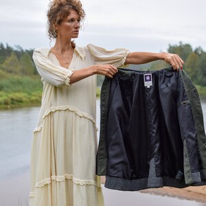 May include: A woman in a white dress holds up a green and black jacket with a purple label. The jacket is unbuttoned and the lining is visible. The woman is standing in front of a river and trees.
